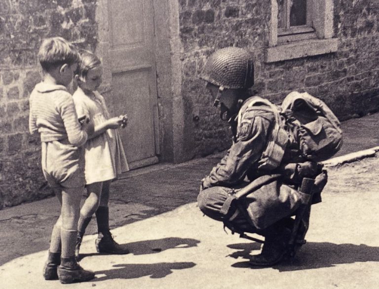 Children from Sainte-Mere-Eglise talk to an 82nd trooper, despite the ongoing battle raging around the town.
